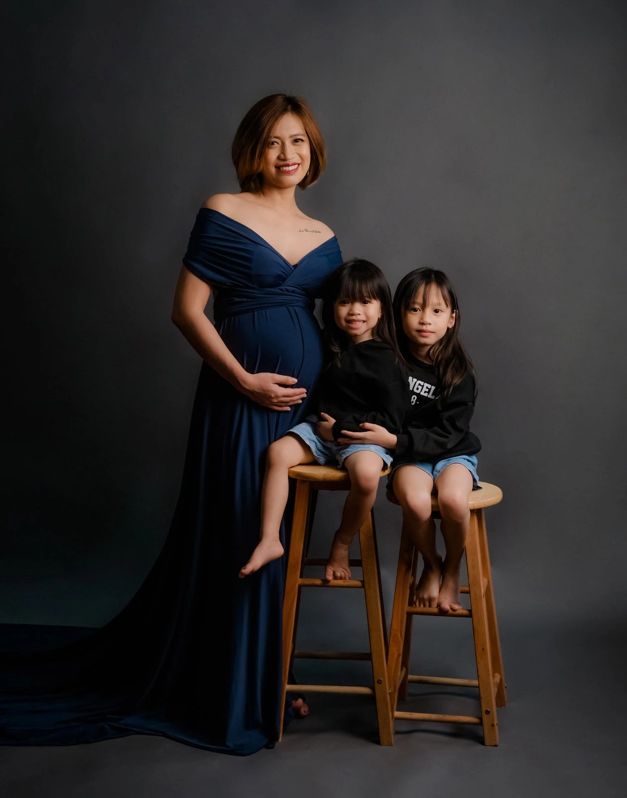 Ces, pregnant, smiling while standing beside her two daughters in a studio portrait.