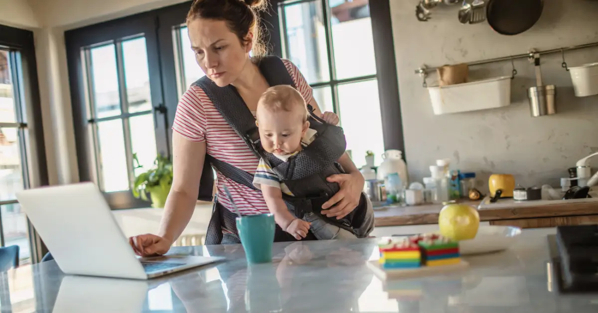 Mother working on a laptop while carrying her baby, representing how mom guilt shows up when balancing work and parenting.