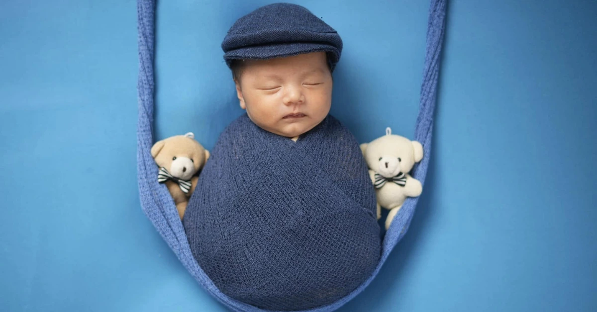 Newborn baby in a blue swaddle with matching cap, sleeping between two teddy bears, representing the idea of rest when baby sleeps.