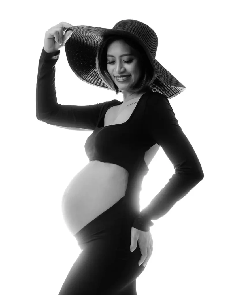 Pregnant woman in a stylish black hat holding her belly during a maternity shoot.